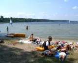 Ludzie odpoczywają na plaży i kąpią się w jeziorze. W tle żaglówki i las. To typowe boszkowo atrakcje w letni dzień.