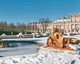 Schneebedeckte Gärten und ein gefrorener Brunnen vor einem Palast in Spanien im Winter. Eine bronzene Statue blickt auf die verschneite Landschaft.