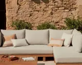 Sofá modular de exterior con cojines en tonos neutros, sobre una mesa de madera. La **decoración estilo natural** se realza con la pared de piedra y la vegetación.