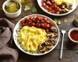 Talerz z puree ziemniaczanym, parówkami i pieczarkami, obok ogórki i sos.