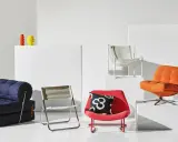 En samling möbler i 70-tals inredning: en blå bäddsoffa, en röd fåtölj med mönstrat kudde, en orange fåtölj och en vit metallstol.