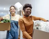 Deux amies souriantes déménagent, portant des cartons remplis d'objets. Une nouvelle aventure commence, peut-être grâce à un **bail solidaire**.