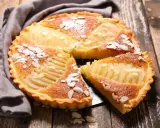 Une délicieuse tarte aux poires, garnie d'amandes effilées et saupoudrée de sucre glace, repose sur un plateau en bois.