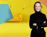 Frau mit Armen verschränkt vor gelbem Hintergrund mit Sofa und Beistelltisch. Inspiration für Renovierungs Serien Deutsch.