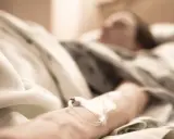 Ręka pacjenta z wenflonem w szpitalnym łóżku, w tle niewyraźna postać.