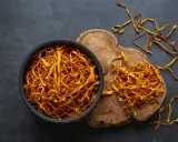 Un bol de cordyceps séchés, connus pour leurs nombreux bienfaits, est posé sur un fond sombre. Quelques brins dorés s'échappent du bol.