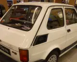 Biały Fiat 126p bis z naklejkami na tylnej szybie, stojący obok czerwonego samochodu.