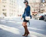 A woman walks down a city street wearing a black dress, a denim jacket, and stylish brown boots.