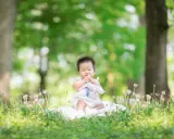 Dolce bimbo di 9 mesi seduto sull'erba, con soffioni e alberi sullo sfondo. Una foto che cattura la sua curiosità.