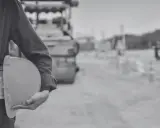 Budowa drogi, pracownik w kamizelce odblaskowej trzyma kask. W tle walec drogowy i znaki ostrzegawcze, symbolizujące wywłaszczenie terenu pod inwestycję.