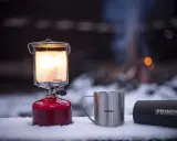 En varm primus lampa lyser upp snön bredvid en mugg och en termos. I bakgrunden syns en lägereld.