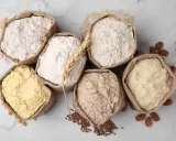 Six sacs en papier remplis de différentes farines, dont une farine de blé CRC. Amandes et grains parsèment le fond marbré.