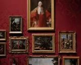 A gallery wall displays classical paintings in ornate frames against a rich red damask background. This collection showcases the best way to invest in art, with portraits and landscapes hinting at historical narratives.