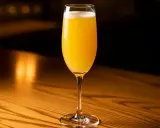 Ett glas med en gyllene mimosa drink, bubblig och uppfriskande, står på ett träbord i varmt ljus.