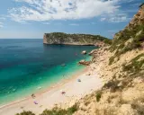 Türkisblaues Wasser trifft auf Kiesstrand in einer malerischen spanischen Atlantikbucht. Felsen und grüne Vegetation säumen die Küste.