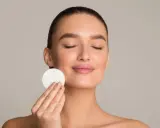 Donna con occhi chiusi applica un dischetto di cotone sul viso, parte della sua routine di skincare viso per una pelle radiosa.