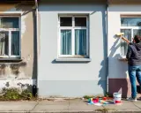 Un homme est en train de refaire un mur extérieur avec un rouleau. Des échantillons de couleurs et un pot de peinture sont posés au sol.