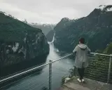 Kobieta podziwia majestatyczny **fjord** z wodospadem i ośnieżonymi szczytami w tle.