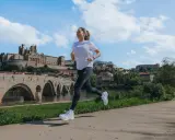 Kobieta biegnie wzdłuż rzeki, mijając zabytkowy most i miasto. Zastanawia się, ile się biegnie 5 km.