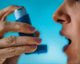 Person nutzt Asthmaspray vor oder nach dem Sport. Die Hand hält das blaue Gerät, der Mund ist geöffnet.