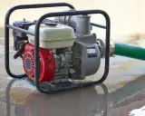 Una motobomba roja y plateada, con una manguera verde, está sumergida en agua turbia, lista para drenar.