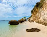 Sonniger Setúbal Strand mit türkisfarbenem Wasser, Felsen und Booten im Hintergrund. Eine kleine Gruppe entspannt sich unter einem Sonnenschirm.