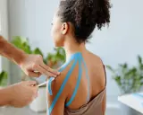 Therapeut legt blaues Kinesio-Tape an Schulter an. Vielleicht ein neues, modernes "Omas Hausmittel gegen Schulterschmerzen"?