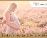 Kobieta w kwiecistej sukience, w 35 tygodniu ciąży, delikatnie dotyka brzucha na tle kwitnącej łąki.