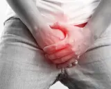 Dłoń obejmuje krocze z czerwonym podświetleniem, sugerując ból lub uraz, jak złamanie prącia.