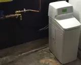 Appareil blanc avec écran vert, connecté à des tuyaux en cuivre. C'est un adoucisseur d'eau potable pour une maison.
