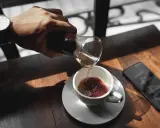 Hand gießt Kaffee in eine Tasse, daneben ein Smartphone mit Uhrzeit.