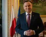 Prezydent Andrzej Duda w garniturze, z polską flagą w tle, czeka na moment, kiedy prezydent podpisze emerytury stażowe.