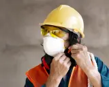 Trabajador ajustando su mascarilla, gafas y epi casco amarillo. Chaleco naranja y protectores auditivos rojos.