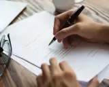 Mano firmando un contrato sencillo de alquiler de vivienda. Gafas y tablet sobre la mesa.