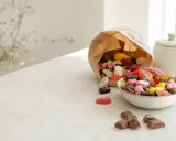 En skål med blandat godis, inklusive geléhallon och chokladbitar, bredvid en papperspåse som svämmar över av fler sötsaker. Perfekt för att temperera choklad till bakning.