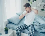 Junger Mann sitzt auf dem Bett, reibt sich den Nacken. Er hat nackenschmerzen nach dem schlafen.