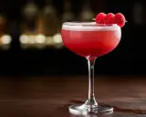 En vackert rosa raspberry sour-cocktail, garnerad med tre färska hallon på en träpinne, står på ett mörkt träbord.