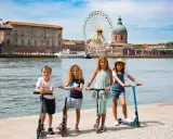 Niños en patinetes junto al río en Toulouse, con la noria y la cúpula de la Basílica de San Sernín de fondo. ¡Un plan genial que hacer en Toulouse con niños!