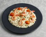Kremowe risotto z krewetkami, pomidorkami koktajlowymi i posiekaną natką pietruszki na czarnym talerzu.