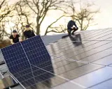 Arbetare installerar solpaneler på ett tak. En av dem, en kvinna, ler glatt medan hon håller i en panel. En annan man hjälper till, och en tredje person sitter på taket. De är en del av teamet för **solverkets alfa**.