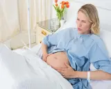 Gravid kvinna i sjukhussäng, med händerna på magen, ser ut att ha värkar. Vattnet har gått och hon väntar på barnet.