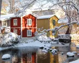 Vinteridyll vid vattnet i destination Gåsvik. Röda och gula hus speglas i det isiga vattnet, omgivna av snötyngda träd.