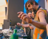 DJ Villa Lobos, con cuffie al collo, mixa musica con passione. Dietro di lui, il pubblico attende il ritmo.