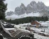 Panorami innevati con il rifugio delle Odle in lontananza, panchine in legno e pini.