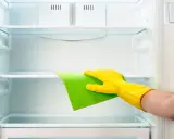 Ręka w żółtej rękawiczce czyści półkę lodówki zieloną ściereczką. Dowiedz się, czym myć lodówkę, by była lśniąca.