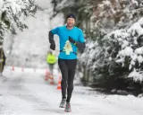 Mann läuft im Winter durch verschneiten Park. Er trägt ein blaues Shirt mit einer Startnummer in Form eines Weihnachtsbaums.