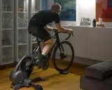 Mężczyzna trenuje na rowerze stacjonarnym z napędem **direct drive**, śledząc wirtualną trasę na laptopie.