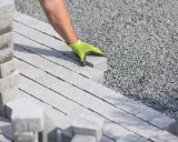 Ręka w zielonej rękawicy układa kostkę brukową na podsypce cementowo piaskowej.