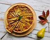 Apetyczna tarta z gruszkami, udekorowana natką pietruszki i skórką cytrynową, prezentuje się na drewnianej desce. Obok leży świeża gruszka i jesienne liście.