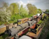 Un cementerio de trenes con efecto tilt shift, donde locomotoras y vagones oxidados parecen miniaturas bajo un cielo nublado.